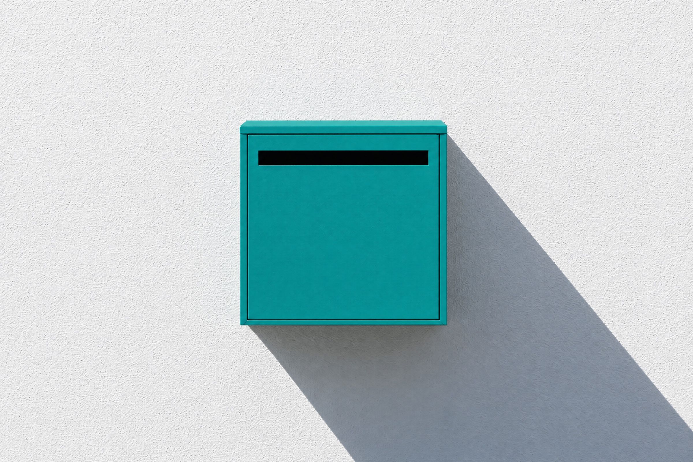An empty teal letterbox on a sunlit white wall, its slot waiting.